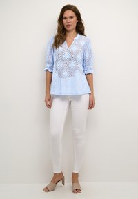 Blusa de encaje azul claro con patrones florales, escote en V y mangas 3/4, combinada con jeans skinny blancos y sandalias de tacón nude.