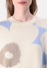 Adīta džemperis ar krēmkrāsas pamatu, ar zilu un brūnu abstraktu rakstu. Apaļa kakla izgriezums un teksturēta auduma vidējais svars.