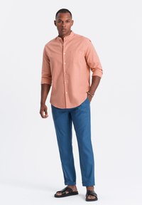 Chemise longue à manches roses pâles avec un col à boutons, assortie à un pantalon bleu et des sandales noires. Comprend une poche poitrine et des manches retroussables.