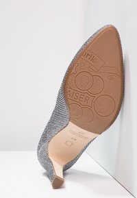 Šedá třpytivá vysoká boty s špičatou špičkou ležící na boku, ukazující embosovanou hnědou podrážku s dekorativními mincovními vzory a textem značky.