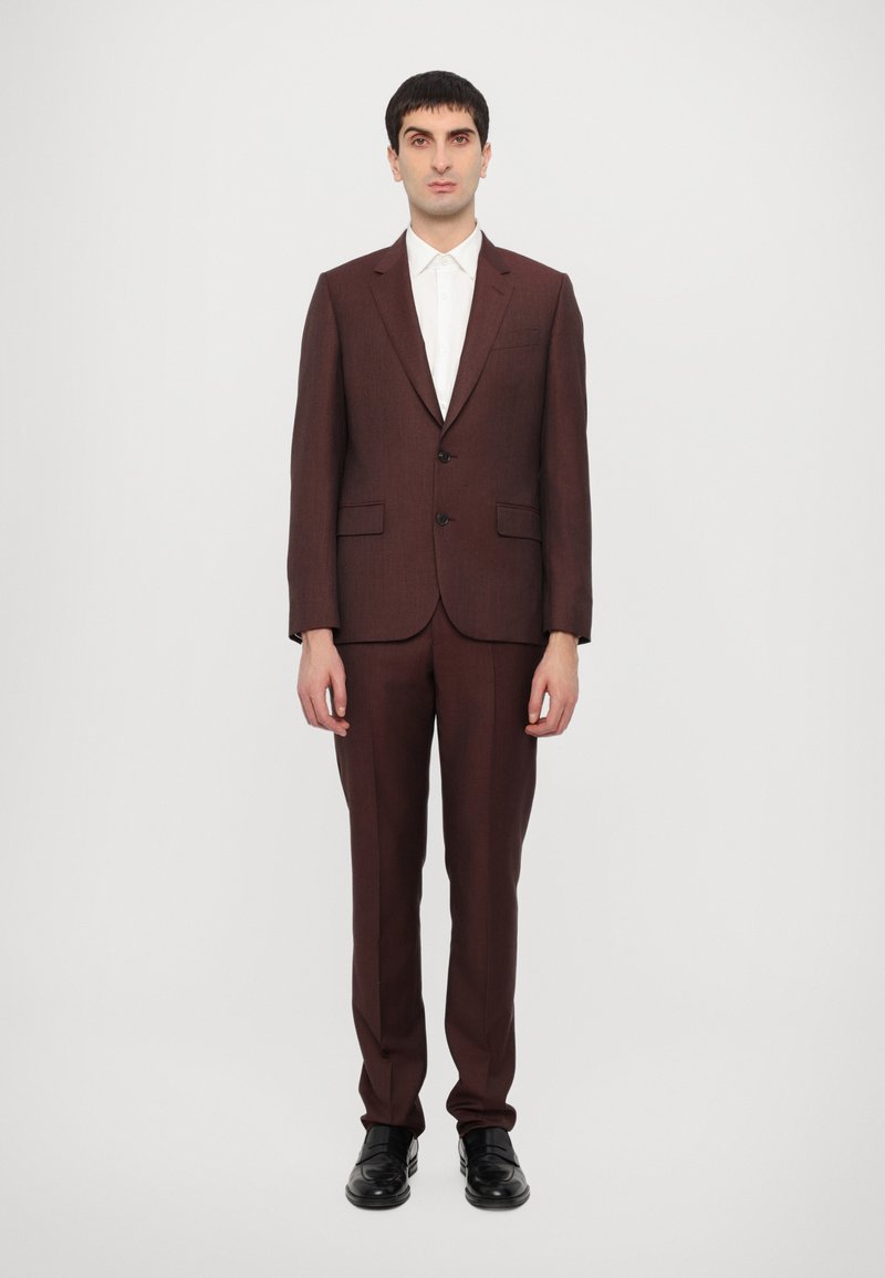 Costume taillé marron foncé avec une texture lisse, conception à un seul bouton, boutons noirs et pantalon assorti. Chemise blanche à col en dessous.