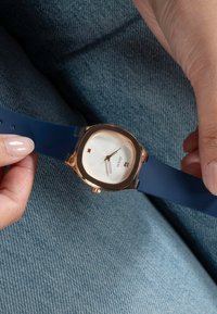 Kvarcóra kerek fehér számlappal, arany részletekkel és kék szilikon szíjjal. Négyzet alakú órajelzővel a 12-es számon és dátumkijelzővel rendelkezik.