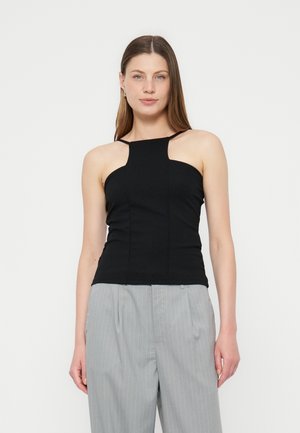 Γυναίκα που φοράει μαύρο αμάνικο τοπ halter και ανοιχτό γκρι παντελόνι με φαρδιά μπατζάκια και λεπτές ρίγες, στέκεται μπροστά σε απλό φόντο.