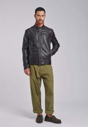 Man staand, gekleed in een zwarte leren jas, olijfgroene omgeslagen broek, bruine schoenen, met één hand in de jaszak tegen een effen achtergrond.