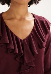 Top burdeos con un profundo escote en V y detalles de volantes verticales en el collar. La tela parece suave con una textura lisa.