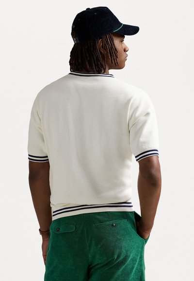 Hvit strikket polo skjorte med marineblåstripede detaljer, kombinert med grønne teksturerte shorts, vist fra baksiden. Caps i mørk farge.