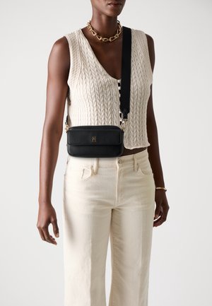 Femme portant un haut sans manches en maille torsadée crème, un pantalon taille haute crème, un sac bandoulière noir, un collier chaîne en or et un bracelet au poignet droit.