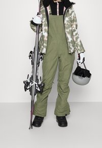 Skibekleidung bestehend aus einer Jacke mit Camouflage-Muster, olivgrünen Latzhosen, schwarzen Skischuhen und einem grauen Helm, mit Skiern, die an der Seite gehalten werden.
