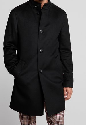 Mann trägt einen schwarzen, knopfgeschlossenen Mantel mittlerer Länge mit den Händen in den Taschen und darunter sichtbaren karierten Hosen.