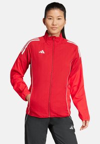 Rote leichte Jacke mit durchgehendem Reißverschluss, weißen Streifen auf den Schultern und seitlichen Taschen. Hergestellt aus glattem, atmungsaktivem Stoff.