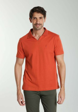 Homme aux cheveux bruns courts portant un polo orange à manches courtes et un pantalon vert foncé, debout devant un fond uni.