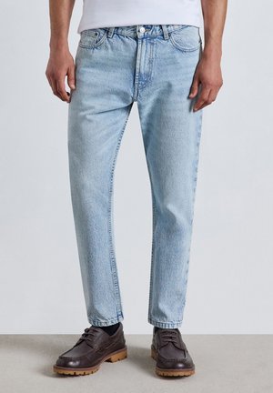 Uomo che indossa jeans azzurri a gamba dritta, scarpe di pelle marrone scuro e una camicia bianca, in piedi su un pavimento grigio.