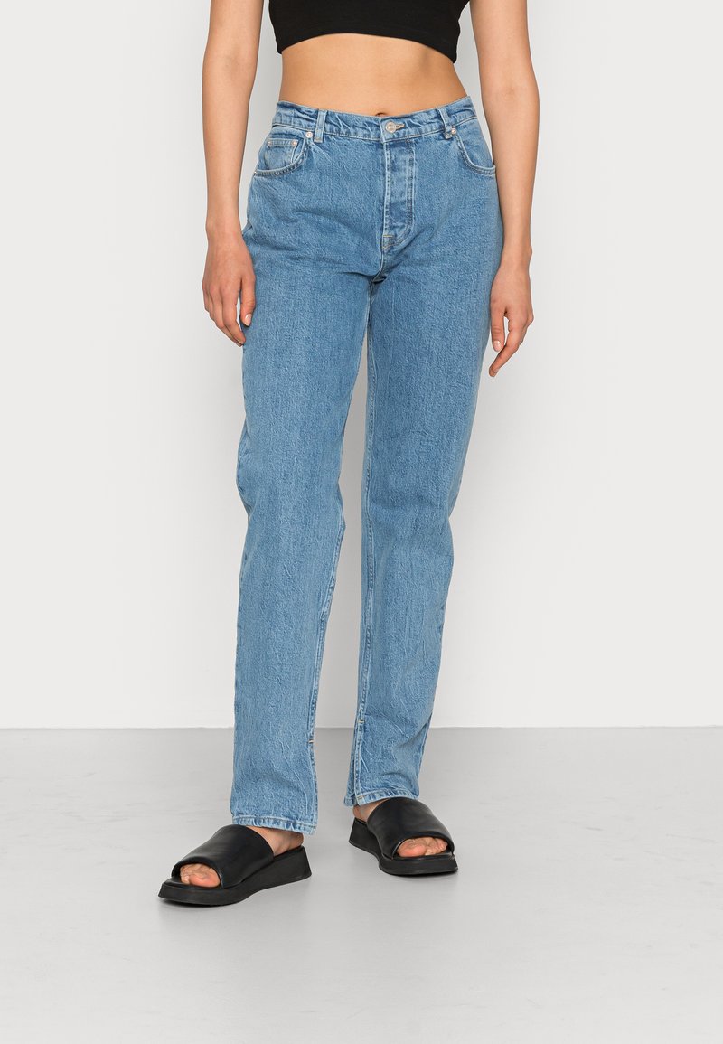 Blue jeans de ganga claros com um corte descontraído, design de cinco bolsos e fecho com botão. Combinados com sandálias pretas de dedo aberto.