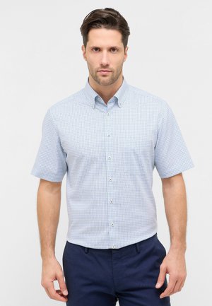 Uomo adulto che indossa una camicia a maniche corte a quadri azzurri con bottoni e pantaloni blu scuro, in piedi contro uno sfondo chiaro e semplice.