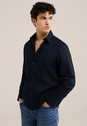 Junger Mann mit lockigem Haar, der ein dunkelblaues Hemd mit Knöpfen und blaue Jeans trägt, die Hände in den Taschen, schaut zur Seite.