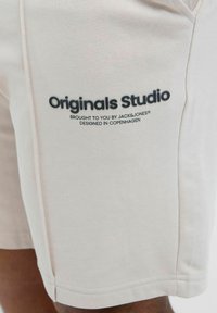 Svijetlosmeđi šortsevi izrađeni od mekog materijala, s patentiranim rubom i plavim logotipom s natpisom "Originals Studio" sa strane.