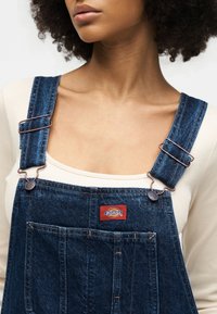 Γυναίκα που φοράει μπεζ μπλουζάκι κάτω από μπλε τζιν ολόσωμες φόρμες Dickies με ρυθμιζόμενους ιμάντες και μπροστινή τσέπη, ορατή από τους ώμους μέχρι το στήθος.