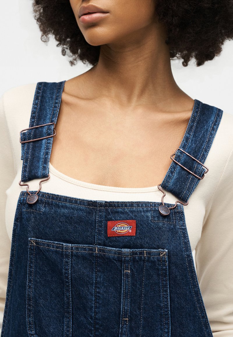 Frau trägt cremefarbenes Oberteil unter blauen Denim-Dickies-Latzhosen mit verstellbaren Trägern und Vordertasche, sichtbar von den Schultern bis zur Brust.