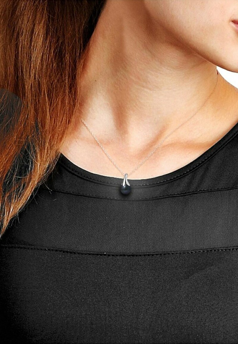 Collier en chaîne argenté avec un pendentif en forme de goutte noire, porté sur un haut noir avec un design texturé et superposé.