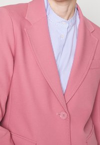Blazer rosa ajustado con solapa de muesca, cierre de un solo botón y detalles de costura sutiles. En el interior presenta una camisa de rayas azul claro.