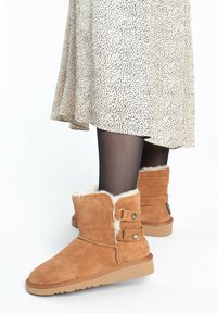 Stivaletti in suede marrone con fodera in shearling, dotati di due cinghie laterali con borchie metalliche e suola in gomma. Abbinati a un vestito a pois.