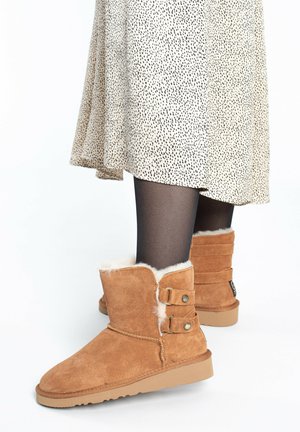 Brown suede ankle boots with shearling lining, featuring two side straps with metal studs and a rubber sole. Paired with a spotted dress.