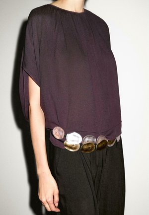 Personne portant un haut ample violet foncé associé à un pantalon noir et une ceinture ornée de grands disques ovales métalliques dorés et argentés.