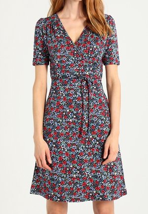 Vrouw draagt een knielange wikkeljurk met korte mouwen, voorzien van een rood, blauw en wit bloemmotief en een strikceintuur in de taille.