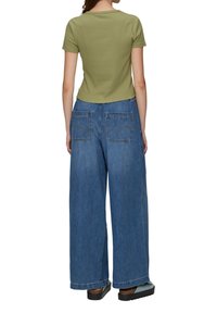 Olijfgroen top met korte mouwen, gecombineerd met wijde spijkerbroek in blauw met twee achterzakken en zichtbare stiksels. Zwarte schoenen.