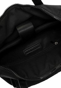 Interior de una bolsa de tela negra con una textura suave, bolsillo con cremallera y etiqueta de marca. Presenta una cremallera robusta y una correa de textil.