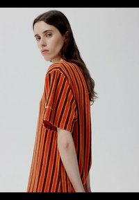 Žena s dugom smeđom kosom koja nosi kratku majicu s vertikalnim narančasto-smeđim prugama, gleda preko ramena na jednobojnoj pozadini.