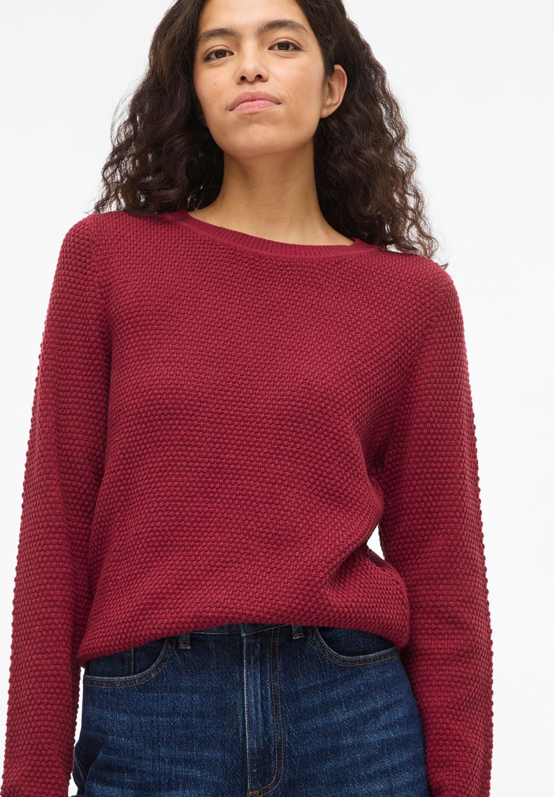 Γυναίκα με σγουρά σκούρα μαλλιά, ντυμένη με κόκκινο, υφασμάτινο πουλόβερ μακρυμάνικο και μπλε τζιν, σε φόντο απλό λευκό.