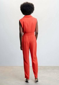 Czerwony bezrękawnik z kołnierzem, taliowany paskiem, z prostymi nogawkami. Gładka tekstura, minimalne szwy, noszony z czarno-białymi szpilkami.