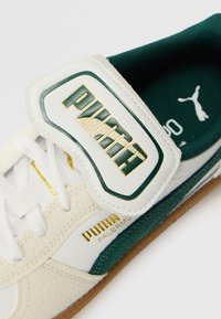 Weißes Leder-Sneaker mit grünen Akzenten und goldenem Logo. Verfügt über einen Klettverschluss und eine strukturierte Oberfläche. Gummisohle mit geripptem Muster.