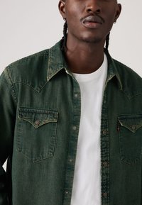 Παντελόνι από σκούρο πράσινο τζιν  με μυτερό γιακά, δύο μπροστινές τσέπες και κουμπιά. Φορεμένο πάνω από λευκό μπλουζάκι, που δείχνει υφασμένο ύφασμα.