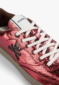 Zapatilla deportiva metálica rosa con acabado texturizado, cordones blancos y un logo negro de calavera y huesos cruzados. Cuenta con un cuello acolchado marrón.