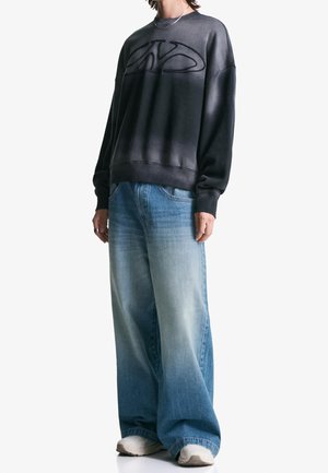 Personne portant un sweat-shirt noir oversize avec un logo en relief, un jean bleu délavé à jambes larges, des baskets blanches et un collier argenté.