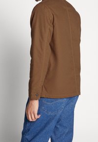 Brown cotton jacket with a straight cut, featuring a visible seam detail and buttoned cuffs, paired with blue denim pants.