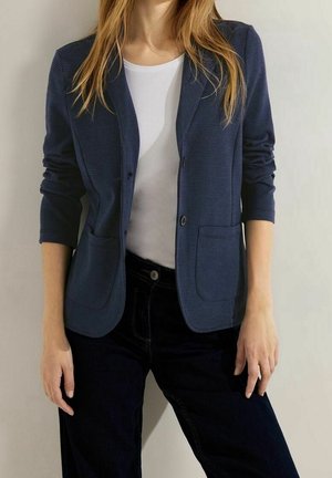 Frau trägt ein marineblaues Sakko mit aufgesetzten Taschen über einem weißen Hemd und schwarzen Hosen, steht mit locker an den Seiten hängenden Armen.