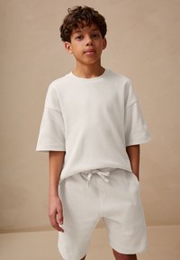 Balti trumpomis rankovėmis tekstūruota marškinėliai ir šortai su elastiniu liemeniu, abu pagaminti iš minkštos medžiagos, demonstruojami ant vaiko priešais paprastą foną.