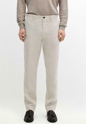 Uomo con pantaloni beige a gamba dritta, camicia marrone chiaro a maniche lunghe e mocassini marroni scuri con nappine, in piedi su sfondo bianco.