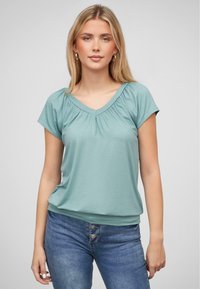 Mintgroene top met korte mouwen en een V-hals, met geplooide details op de schouders. Zachte stof met een ontspannen pasvorm, gecombineerd met denim.