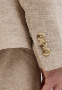 Manga de blazer de lino beige con tres botones beige, con un patrón texturizado sutil y costuras visibles a lo largo del puño.