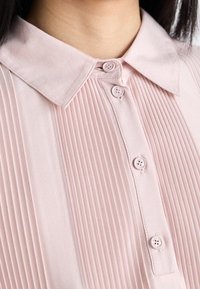 Ljusrosa blus med en veckad design, med krage och knappstängning framtill. Slät textur med små matchande knappar.
