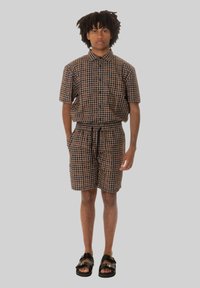 Cameriera a maniche corte marrone e pantaloni corti coordinati con un motivo a pois. Indossata con sandali neri. Il tessuto sembra leggero e traspirante.