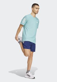 Camiseta deportiva azul claro con diseño de tres rayas blancas en las mangas, combinada con pantalones cortos azul marino. Zapatillas deportivas con detalles en negro.
