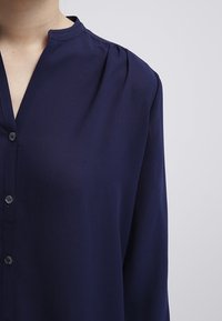 Blusa blu navy in tessuto leggero, dotata di colletto alla cinese, chiusura con bottoni e dettagli arricciati sulle spalle.