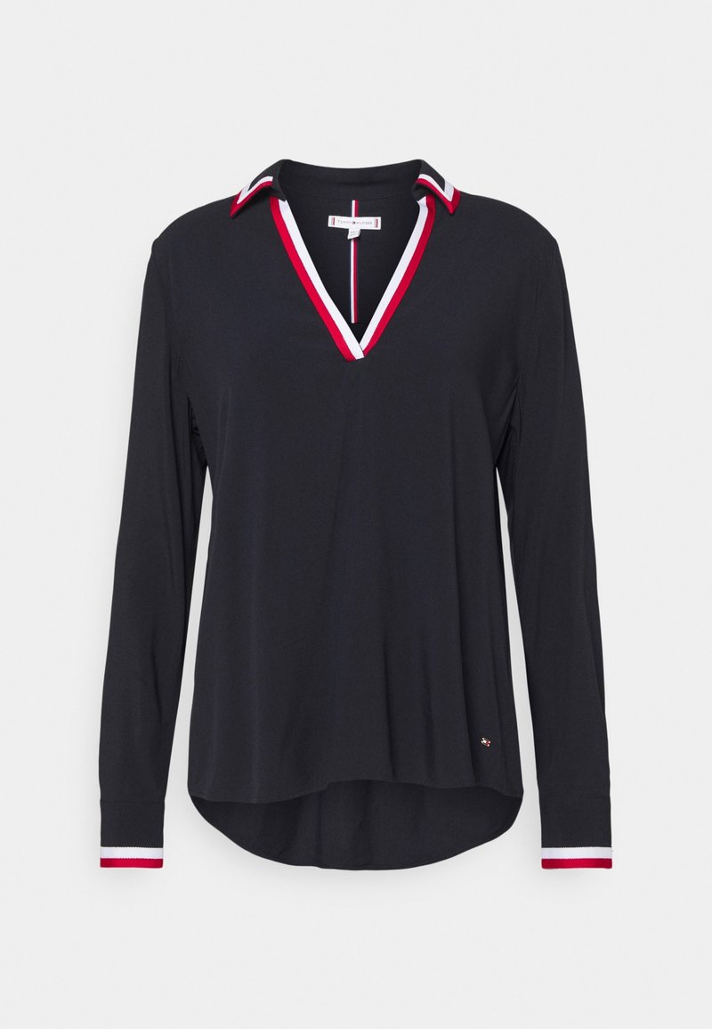 Chemise noire à manches longues avec col en V orné de rayures rouges et blanches, poignets assortis rayés, et ourlet arrière légèrement plus long.