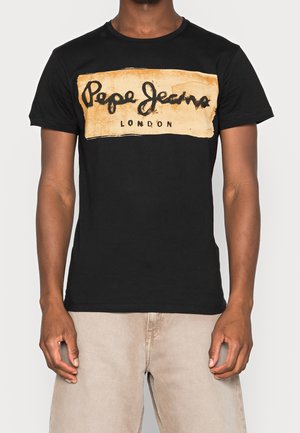 Mann trägt ein schwarzes kurzärmeliges T-Shirt mit einem beigen rechteckigen Logo, auf dem "Pepe Jeans London" steht, und beige Hosen.