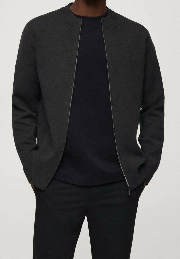 Homme portant une veste zippée gris foncé par-dessus un t-shirt noir, les mains dans les poches, debout devant un fond clair uni.